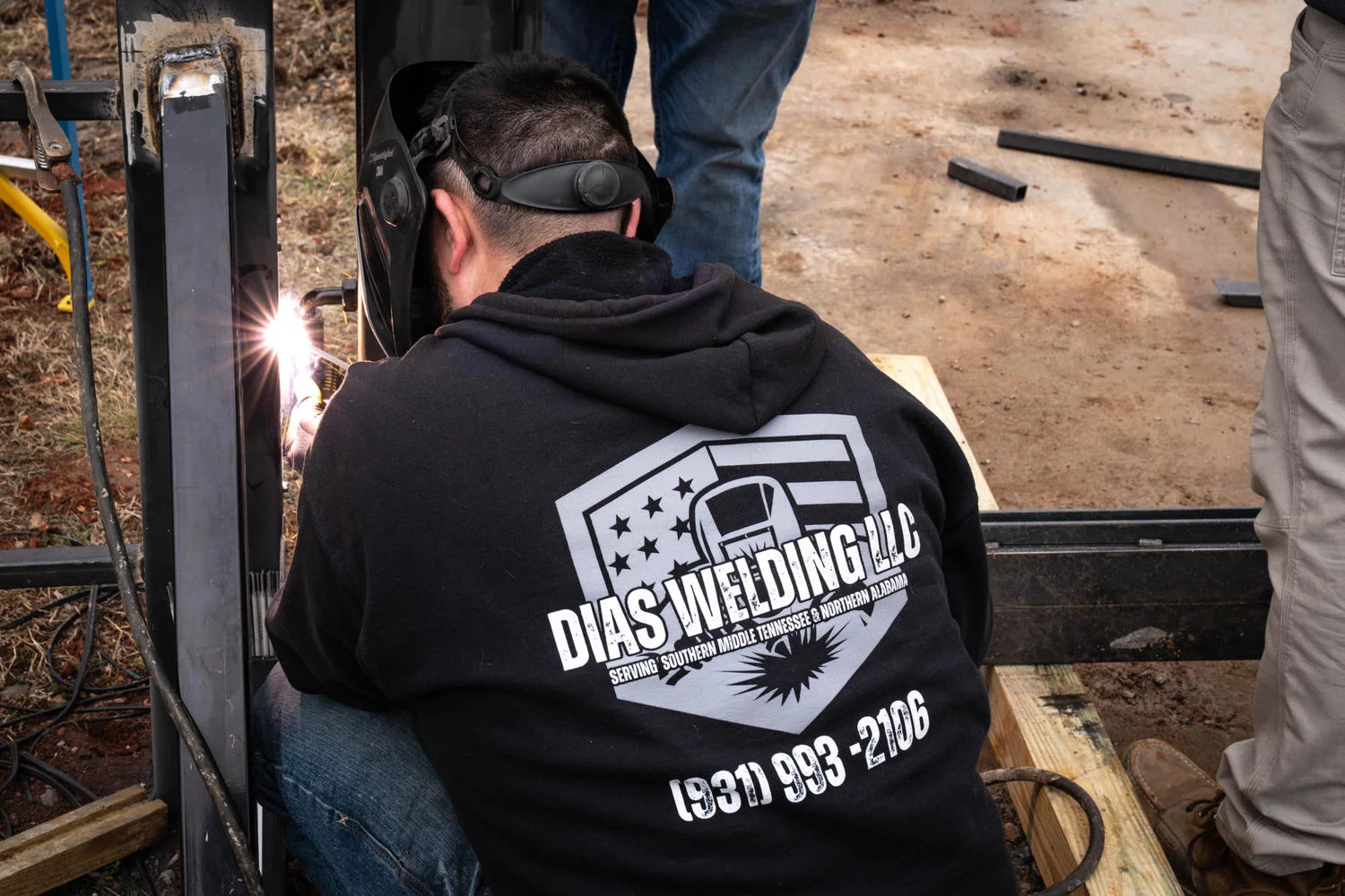 A person in a black hoodie and welding mask is welding a metal structure with bright sparks flying.